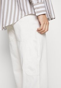 Camisa a rayas en gris claro y blanco, con dobladillo curvado y puños abotonados, combinada con pantalones anchos blancos con bolsillos visibles.
