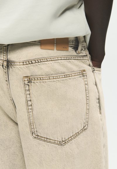 Άτομο που φοράει ανοιχτό μπεζ τζιν από denim με ορατή πίσω τσέπη και ετικέτα από δέρμα ταμπά ραμμένη κοντά στη μέση.