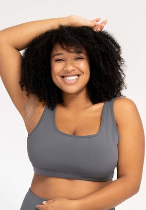 Woman with curly hair smiling, wearing a gray sports bra, with one arm raised and the other resting on her waist.