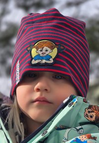 Raidallinen pipo tummansinisenä ja punaisena, jossa on brodeerattu tyttökuva; lapsi pukeutuneena kuviolliseen vaaleanvihreään takkiin, jossa on sarjakuvakuvastoa.