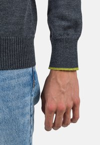 Grauer Strickpullover mit gerippten Bündchen; das innere Bündchen hat einen leuchtend grünen Akzent. Getragen mit hellblauer Jeans, wobei ein Teil der Hand sichtbar ist.
