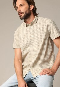 Chemise beige à manches courtes avec un motif de petits points noirs et bleus, dotée d'un col boutonné et de boutons blancs. Texture de tissu douce.