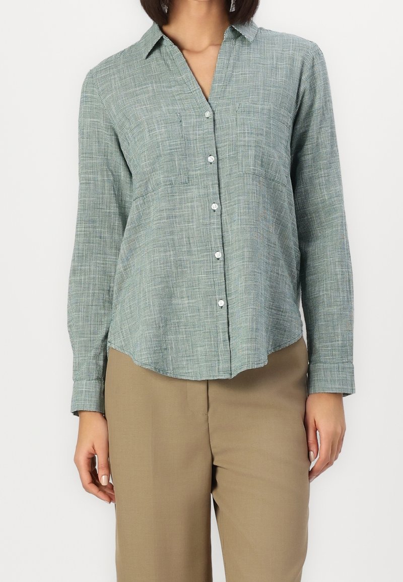 Femme portant une chemise à carreaux vert clair à manches longues avec des boutons et un pantalon beige, debout devant un fond blanc uni.