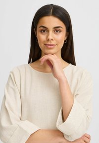 Top en lin beige avec un décolleté bateau et des manches roulées. Texture lisse avec un motif tissé subtil. Le mannequin pose la main sur le menton.