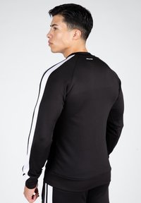 Sweatshirt de sport noir avec des rayures blanches sur les épaules, design ajusté, texture lisse, poignets côtelés et logo au niveau de l'encolure arrière.