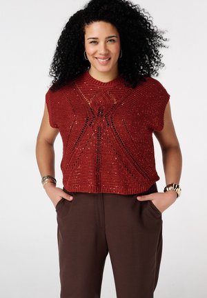 Femme aux cheveux bouclés portant un top rouge en maille sans manches avec des motifs et un pantalon marron, souriante, les mains dans les poches.