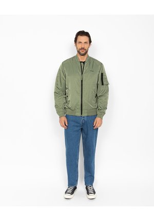 Homme debout portant une veste bomber vert olive, un jean bleu et des baskets noires sur un fond blanc.