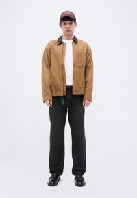 Νεαρός άνδρας που φοράει καφέ μπουφάν, λευκό T-shirt, σκουρόχρωμο τζιν, μαύρα παπούτσια και μπορντό καπέλο, στέκεται μπροστά σε απλό λευκό φόντο.