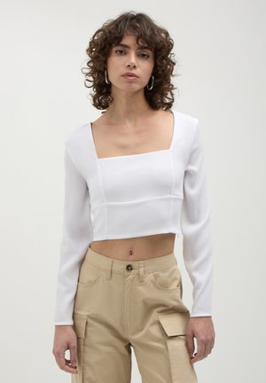 Top bianco corto con maniche lunghe e scollatura quadrata. Tessuto liscio con dettagli delle cuciture. Abbinato a pantaloni cargo beige con tasche.