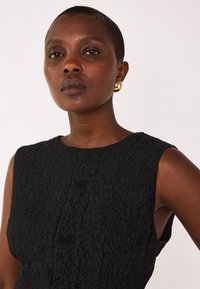 Schwarzes ärmelloses Kleid aus strukturiertem Stoff, mit Rundhalsausschnitt und seitlichen Ausschnitten. Goldene runde Ohrringe setzen Akzente zu dem Look.