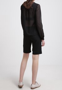 Donna con una blusa nera trasparente a maniche lunghe, pantaloni corti neri fino al ginocchio e scarpe beige con lacci, in piedi rivolta verso la parte opposta su uno sfondo neutro.