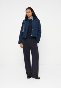 Casaco acolchoado azul-marinho com colarinho, bolso no peito e fecho de correr prateado; combinado com um suéter azul-marinho de malha trançada e calças de perna larga.