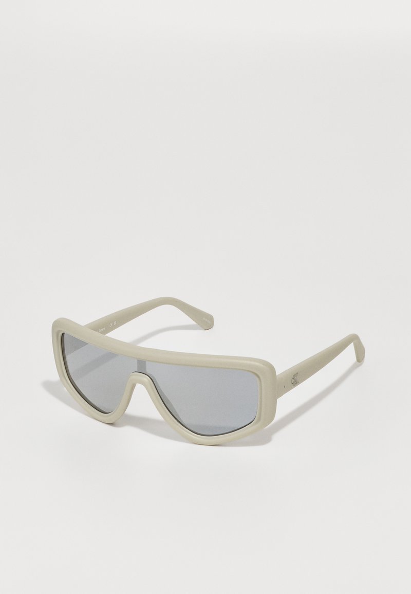 Beige oversized sunglasses with a thick, matte frame and grey tinted lenses. The lenses are angular with a bold design, featuring straight temples.