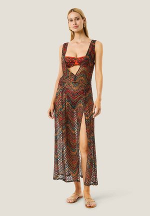 Donna che indossa un vestito lungo senza maniche con motivo, apertura frontale, spacco alto sulla coscia e sandali aperti, in piedi su sfondo semplice.