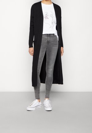 Person trägt ein weißes T-Shirt mit Grafik, langen schwarzen Cardigan, graue Skinny-Jeans und weiße Plateausneaker und steht vor einem schlichten Hintergrund.