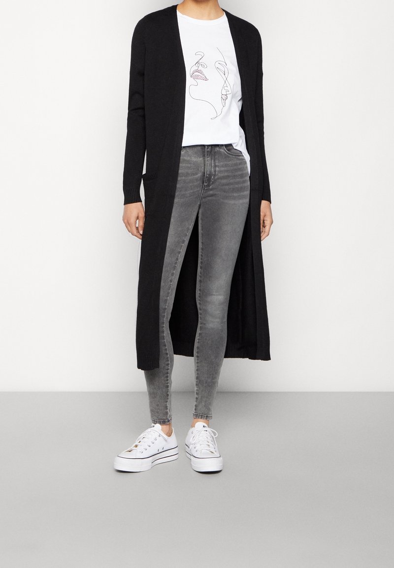 Personne portant un t-shirt blanc à motif graphique, un long cardigan noir, un jean skinny gris et des baskets blanches à plateforme, debout devant un fond uni.