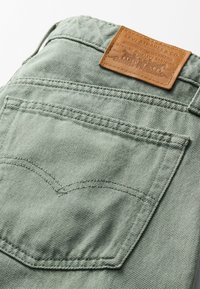 Zeleni denim material z našitim zadnjim žepom, ki vsebuje rjavo usnjeno etiketo z vtisnjenim tekstom in grafiko, ki označujeta znamko in kakovost.