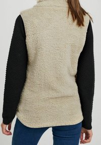 Beige Fleece-Weste mit hohem Kragen und strukturierten schwarzen Ärmeln, ausgestattet mit einer glatten Außenschicht und weichem Innenfutter. Gerundeter Saum.