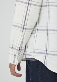 Chemise à carreaux blanche et lavande, en tissu texturé, coupe décontractée, avec des manches retroussées et un ourlet incurvé pour faciliter les mouvements.