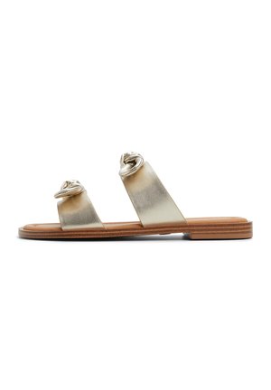 Platta sandaler med två metalliska guldfärgad remmar som har knutdetaljer, beige fotbädd och en texturerad sula. Minimalistisk design, öppen tå-stil.