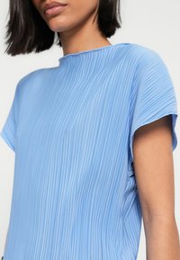 Ανοιχτό μπλε πλισέ τοπ με υψηλό λαιμό, κοντά μανίκια και απαλή υφή. Διαθέτει μαλακό ύφασμα με κάθετα ραβδώσεις.
