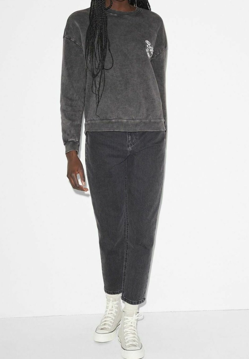 Femme portant un sweat-shirt noir délavé, une jupe midi en jean noire et des baskets blanches montantes sur fond blanc uni.