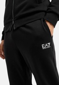Pantalones deportivos negros con una textura suave, que presentan un prominente logo blanco de EA7 en el muslo y un bolsillo con cremallera en la cintura.
