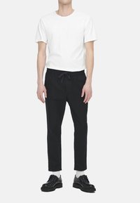 Weißes Rundhals-T-Shirt aus leichtem Stoff, kombiniert mit schwarzen Jogginghosen in lockerer Passform, ergänzt durch schwarze Schuhe und weiße Socken.