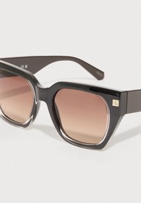 Óculos de sol retangulares pretos com lentes castanhas graduadas, armações grossas, acabamento lustroso e detalhe do logótipo dourado CK na lateral.