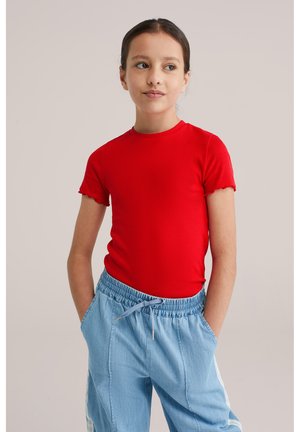 Djevojka s tamnom kosom u crvenoj majici kratkih rukava i svijetloplavim hlačama s elastičnim strukom stoji s rukama u džepovima i gleda desno.