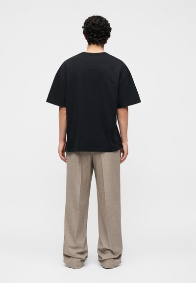 Άτομο που στέκεται με την πλάτη, φοράει χαλαρό μαύρο κοντομάνικο πουκάμισο και φαρδιά μπεζ παντελόνια σε απλό φόντο.