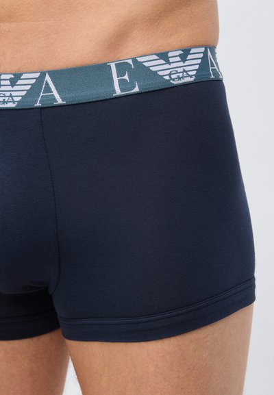 Κοντινό πλάνο ενός ατόμου που φορά μπλε ναυτικό boxer briefs με σκούρο τιρκουάζ ζώνη μέσης που φέρει λευκά γράμματα και λογότυπα αετού.