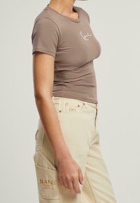 Brązowy dopasowany t-shirt z krótkim rękawem i białym logo. W połączeniu z beżowymi spodniami cargo z przednimi kieszeniami i kremowymi szwami.