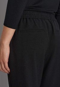 Zwarte broek met een gladde textuur, voorzien van een elastische tailleband en een discreet achterzakdetail, zijwaarts getoond met een hand op de stof.