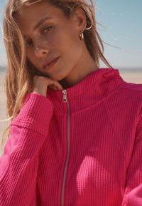 Veste zippée rose vif en tissu texturé côtelé. Caractérisée par un col montant et une fermeture éclair argentée, mettant en valeur une coupe ajustée.