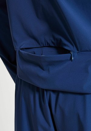 Nærbilde av et marineblått jakkelomme med glidelås delvis åpen på et plagg i matchende stoff.