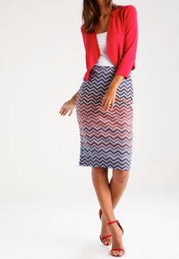 Rode cardigan over een witte tanktop, gecombineerd met een rok met een zigzagpatroon in rood, wit en blauw. De outfit omvat rode sandalen.