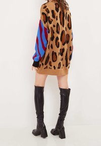 Stickad klänning med leopardmönster på en beige bakgrund och blå och röda randiga ärmar. Svarta ribbade muddar och nederkant. Bruna stövlar över knäna.