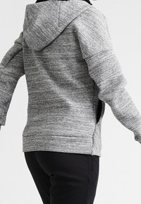 Sudadera gris con un diseño texturizado, que presenta una gran capucha, hombros caídos, aberturas laterales y dobladillo acanalado, combinada con pantalones negros.