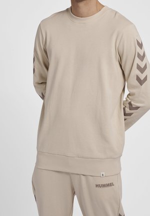 Homme portant un sweat-shirt beige Hummel et un pantalon assorti avec des logos chevron sur les manches et le nom de la marque sur le pantalon.