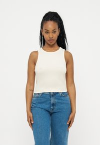 Γυναίκα με μακριά πλεξούδα, φορώντας λευκό μπλουζάκι χωρίς μανίκια και μπλε τζιν, στέκεται μπροστά σε ένα απλό λευκό φόντο.