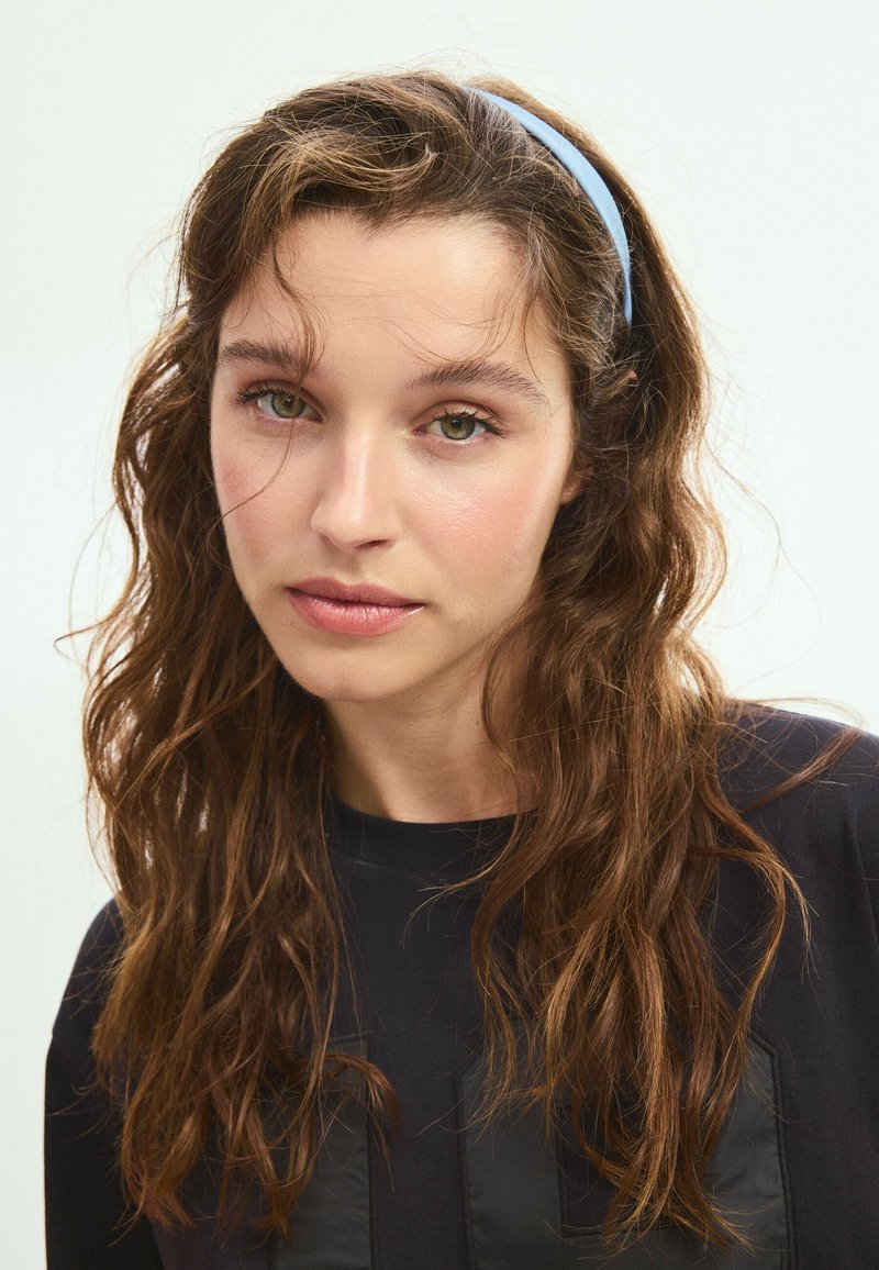 Jeune femme aux cheveux bruns ondulés, portant un bandeau bleu et un haut noir, regardant directement la caméra avec une expression neutre.