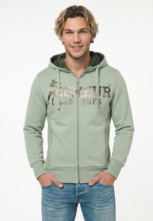 Giovane uomo sorridente che indossa una felpa verde chiaro con cerniera e testo "Boxeur des Rues" e jeans blu, mani nelle tasche, su sfondo bianco.