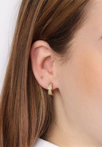 Boucles d'oreilles en or avec une surface texturée et des pierres transparentes incrustées. Petite boucle d'oreille en or portée dans le haut du lobe. Fond de peau lisse.