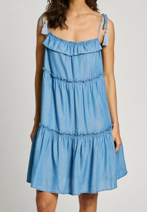 Kobieta w jasnoniebieskiej sukience dżinsowej bez rękawów z falbanami i wiązanymi na ramionach paskami z frędzlami.
