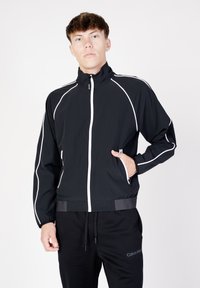 Giacca nera con cerniera realizzata in tessuto leggero, con piping bianco, colletto alto e tasche laterali. Indossata sopra pantaloni della tuta neri.