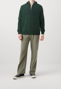 Sudadera con capucha verde con cremallera y un gran logo de "GAP" en navy. Combinada con pantalones holgados de color oliva claro y zapatillas negras.
