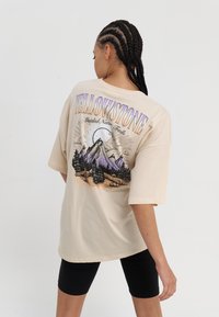 Beige oversized t-skjorte med grafikk av fjell, trær og sol, tekst "YELLOWSTONE," korte ermer, glatt bomullstoff.