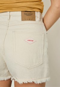 Beigu denimu šorti ar plīša apakšmalu, ar sarkanu dimanta logotipa uzšuvumu aizmugurējā kabatā un brūnu etiķeti jostas vietā.