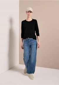 Černá dlouhá košile, světle modré široké džíny a béžová kšiltovka. Model má na sobě bílé tenisky a stojí před neutrálním pozadím.